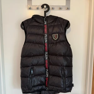 Svart dunväst från Plein Sport - Snygg svart dunväst från Plein Sport med huva och dragkedja framtill. Västen har två fickor med dragkedja och ett emblem med tiger på bröstet. Märkeslogga längs dragkedjan i vitt och rött. Perfekt för lager-på-lager-stil.