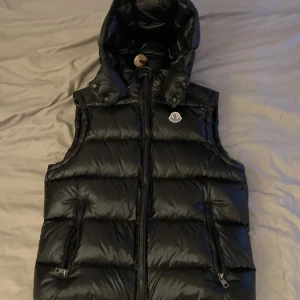 Svart dunväst från Moncler med huva - Säljer en svart dunväst från Moncler med avtagbar huva och dragkedja framtill. Västen har två fickor med dragkedja och klassisk Moncler-logga på bröstet. Perfekt för lager-på-lager under kyliga dagar. Storlek S men passar även M