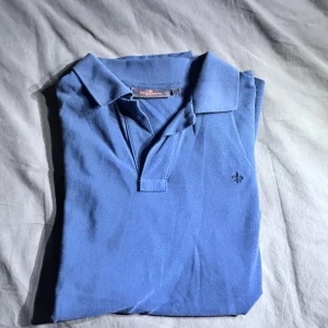 Blå pikétröja från Morris - Säljer en klassisk blå pikétröja från Morris med liten broderad logga på bröstet. Tröjan har krage och korta ärmar, perfekt för en stilren och avslappnad look.