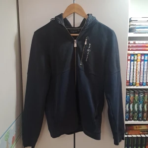 Svart/mörkblå ziphoodiefrån Sail Racing - Svart hoodiejacka från Sail Racing med dragkedja och huva. Jackan har en diskret logga på bröstet och en ficka med dragkedja. Perfekt för en avslappnad stil och enkel att matcha med det mesta. Pris kan förhandlas vid snabbt köp. Självklart äkta!