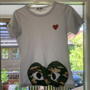 Vit t-shirt från Comme des Garçons Play med camo-detalj - Vit t-shirt med det klassiska röda hjärtat på bröstet och en stor camo-mönstrad hjärtdetalj med ögon längst ner. Enkel och stilren design som sticker ut med coola detaljer.