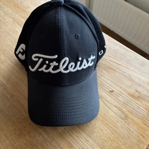 Svart keps från Titleist - Snygg svart keps från Titleist med vit broderad logga framtill. Kepsen har böjd skärm och mesh på sidorna för extra ventilation. Perfekt för sportiga tillfällen eller en casual look.