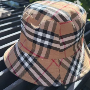  Burberry hat - Säljer en klassisk bucket hat från Burberry i deras ikoniska beigea rutiga mönster med svarta, vita och röda detaljer. Hatten har en mjuk form och är tillverkad i bomull. Perfekt accessoar för att lyfta din stil.