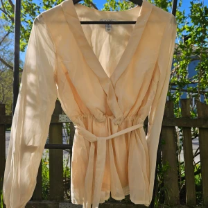 Beige omlottblus med knytband - Säljer en luftig beige omlottblus med långa ärmar och knytband i midjan. Blusen har v-ringning och är perfekt för vår och sommar. Tunn och skön i materialet.