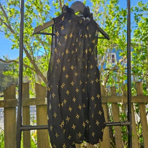 Svart mesh topp med guldiga detaljer  - Säljer en svart ärmlös topp med guldigt geometriskt mönster och hög krage. Toppen har en luftig passform och är perfekt för varma dagar. Stängs med knappar i nacken och har en lätt, flowy känsla.