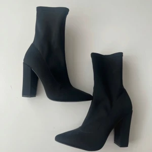 Svarta boots med klack och dragkedja - Säljer ett par svarta boots med hög klack och spetsig tå. Skorna har dragkedja på insidan och är tillverkade i ett stretchigt syntetmaterial som sitter tight runt vristen. 