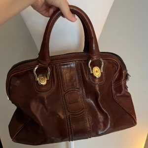 Brun handväska  - Säljer en snygg brun handväska i skinn med två stadiga handtag och guldfärgade metalldetaljer. Kommer med ett band