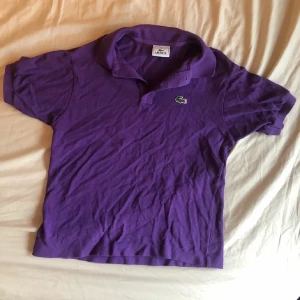 Lila pikétröja från Lacoste - Säljer en klassisk lila pikétröja från Lacoste med den ikoniska gröna krokodilloggan på bröstet. Tröjan har korta ärmar, krage och knäppning framtill. Perfekt för en sportig och avslappnad stil.