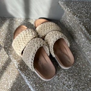 Beige sandaler med grovstickade band - Säljer ett par snygga sandaler  med två breda, grovstickade band i beige färg. Sulan är platt och innersulan är mjuk och bekväm. Perfekta för varma dagar och ger en avslappnad men trendig look.