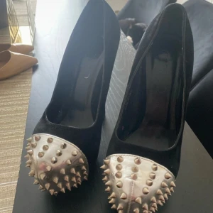 Svarta pumps med silvriga nitar - Säljer ett par svarta pumps med klack och rund tå. Skorna har en unik detalj med silvriga metallhättor framtill, täckta av spetsiga nitar. Materialet är sammet och metall, vilket ger en cool kontrast. Perfekta för dig som vill sticka ut med din stil.