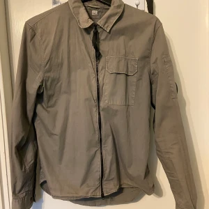 Grå overshirt från CP Company  - Säljer en riktigt snygg övershirt ifrån CP company. Säljer den då den är för liten för mig. Den är tunn och passar perfekt till sommaren och våren att ha utomhus. Tyvärr är det en liten färg fel på kragen (se bild 4).