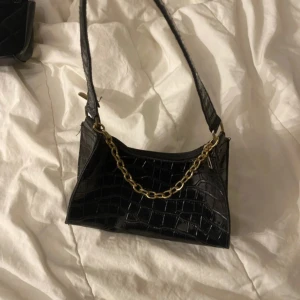 Svart axelväska med kedjedetalj - Snygg svart axelväska i croco-mönstrat syntetmaterial med guldfärgad kedja som detalj. Väskan har dragkedja upptill och ett stilrent, modernt utseende. Perfekt för att lyfta din outfit med en trendig accessoar.