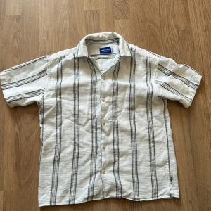 Vit och grå randig skjorta från Jack & Jones - Kortärmad skjorta från Jack & Jones i vit med gråa vertikala ränder. Skjortan har klassisk krage och knäppning framtill. Perfekt för varma dagar och enkel att matcha med jeans eller shorts.