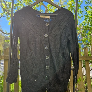 Svart kofta med knappar - Svart kofta med långa ärmar och stora knappar framtill. Koftan har en rund halsringning och är ribbad upptill. Perfekt att slänga över en topp när det blir kyligt.