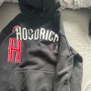 Svart hoodie från Hoodrich med broderade detaljer - Säljer en svart hoodie från Hoodrich med vit logga och stor röd broderad H på framsidan. Tröjan har huva med dragsko, känguruficka och broderade patches. Perfekt för dig som gillar streetwear.