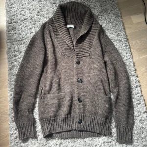 Brun stickad kofta från Bläck - Mysig brun stickad kofta från Bläck med bred krage och knappar framtill. Koftan har långa ärmar, två fickor och ett matchande bälte som kan knytas i midjan. Perfekt för lager-på-lager och chill höst- eller vinterstil.