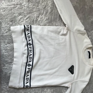 Vit långärmad tröja från Prada - Snygg vit långärmad tröja från Prada med svart logotyp på bröstet och coolt band med Prada-tryck längs nederkanten. Tröjan har rund hals och är tillverkad i mjuk bomull som känns skön mot huden. Perfekt för dig som gillar stilrena designer och exklusiva märken.
