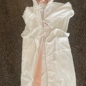 Vit morgonrock med huva Polarn O. Pyret - Mysig vit morgonrock från Polarn O. Pyret i storlek 146/152. Den har huva, två fickor och ett bälte i midjan. Insidan av huvan är ljusrosa och materialet är supermjukt. Perfekt för chilliga dagar när du vill vara extra bekväm.