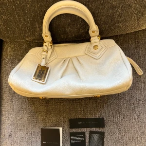  Marc by Marc Jacobs väska  - Snygg vit handväska från Marc by Marc Jacobs i äkta skinn med guldfärgade metalldetaljer och dubbla handtag. Väskan har en klassisk form, dekorativ loggoplatta och räfflad struktur i materialet. Perfekt för dig som vill ha en stilren och tidlös accessoar.