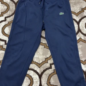 Blå mjukisbyxor från Lacoste - Mörkblå mjukisbyxor från Lacoste med klassisk krokodillogga på ena benet. Byxorna har dragsko i midjan och är tillverkade i mjukt bomullsmaterial. Perfekta för chill dagar eller när du vill ha en sportig look.