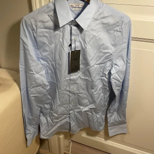 Ljusblå skjorta från Mango XS - Säljer ännu en helt ny ljusblå skjorta från Mango i super slim fit. Klassisk krage, knäppning framtill och långa ärmar. Skjortan är i bomull och passar dig som vill ha en tajt passform.