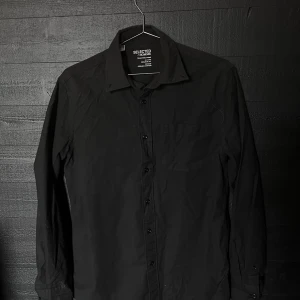 Svart skjorta (Selected Homme) - Svart skjorta från Selected Homme i regular fit med klassisk krage och bröstficka. Tillverkad i ekologisk bomull för en skön känsla. Långärmad modell med knappar framtill.