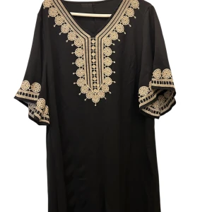 Svart långklänning med broderi - Svart långklänning med v-ringning och unika beiga broderier längs halsringningen och ärmarna. Klänningen har vida, korta ärmar och ett löst, bekvämt fall. Perfekt för dig som gillar detaljer och en bohemisk vibe. Funkar för storlek M och L