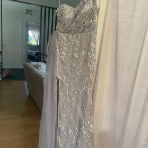 Lång silverfärgad paljettklänning - Säljer en magisk lång klänning i silver med glittriga paljetter och pärlor över hela tyget. Klänningen har axelbandslös design, svepande tyg och vacker slits. Perfekt för bal eller fest när du vill glänsa extra mycket. Materialet är mesh med paljett- och pärldetaljer. Använd endast en gång. Mått: Längd från bröst: 135 cm