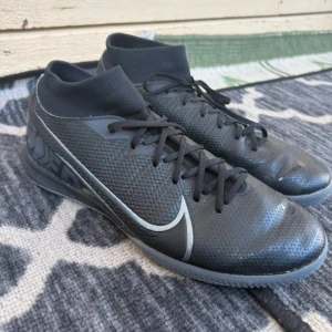 Svarta Nike Mercurial fotbollsskor - Snygga svarta Nike Mercurial fotbollsskor med högt skaft och snörning. Skorna har en strumpliknande passform och är tillverkade i syntetmaterial med en platt, mönstrad sula för bra grepp på inomhusplan. Perfekta för dig som vill ha stilrena och sportiga skor på planen.