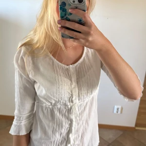 Vit blus med knappar och knyt - Supersöt vit blus med knappar framtill, dekorativa sömmar och knyt under bysten. Trekvartsärm och lätt, luftig bomullskänsla. Perfekt för dig som gillar romantisk och enkel stil.