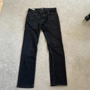 Svarta jeans Jack & Jones Clark 30/34 - Svarta jeans från Jack & Jones, modell Clark Regular. Klassisk femficksdesign med raka ben och normal passform. Jeansen är tillverkade i bomull och har diskreta detaljer som metallknapp. Perfekta för en clean och stilren look.