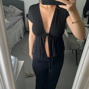 Svart omlottopp med knyt fram - Säljer en svart omlottopp med djup v-ringning och knyt framtill. Toppen har korta ärmar och en lös passform.