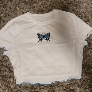 Vit croppad topp med blå fjäril SHEIN - Superfin vit croppad topp från SHEIN med blå fjäril på bröstet. Toppen har korta ärmar och vågiga blå kanter längs ärmar och nederkant. Materialet är en mix av bomull och polyester, vilket gör den mjuk och skön.