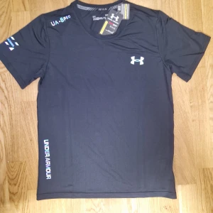 Svart tränings-tshirt Under Armour - Svart t-shirt från Under Armour med logga på bröstet och tryck på ärm och sida. Gjord i ett lätt och snabbtorkande material som passar perfekt för träning. Klassisk passform med korta ärmar och rund hals. Snygg och sportig vibe.