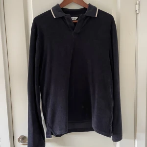 Mörkblå pikétröja från East West - Snygg mörkblå långärmad pikétröja från East West med klassisk krage och vit kantdetalj. Tröjan har en relaxed passform och är tillverkad i mjukt material som känns skönt mot huden. Perfekt för dig som gillar stilren och enkel design.