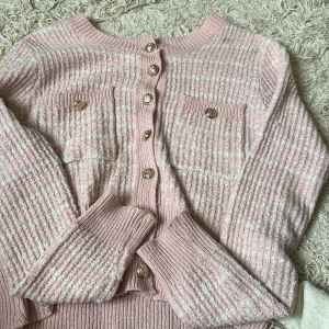 Rosa kofta med guldknappar - Supersöt rosa kofta med vita ränder och snygga guldknappar framtill. Koftan har två bröstfickor med dekorativa knappar och är ribbstickad vid ärmslut och nederkant. Perfekt för lager-på-lager och ger en chic vibe till din outfit. Köpt tidigare ifrån Plick för 300, Xs-s