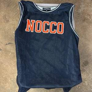 Blått basketlinne NOCCO Official Apparel - Sportig blå meshtröja från Official Apparel med NOCCO-tryck i orange och vitt framtill. Ärmlös modell med vita ränder längs hals och ärmöppningar. Perfekt för träning eller som statement-plagg på sommaren.