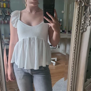 Vit volangtopp från 157, XS - Supersöt vit topp från 157 i storlek XS. Modellen har volangärmar, v-ringning och en flowy, rynkad nederdel. Tyget är lätt strukturerat och toppen är ärmlös med en luftig känsla. Perfekt för en fräsch och somrig look!