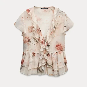 Blommig volangblus från Zara - Supersöt blommig blus från Zara i ljusa toner med volanger längs v-ringningen och nertill. Blusen har korta ärmar och knyt framtill. Endast använd en gång.