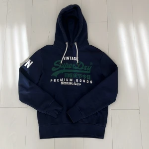 Marinblå Superdry - Mörkblå hoodie från Superdry med grönt och vitt tryck på bröstet och texten 'JPN' på ärmen. Klassisk känguruficka, vita snören i huvan och mjukt material. Perfekt för dig som gillar streetwear och vill ha en snygg, avslappnad look.