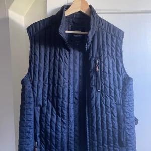 Mörkblå quiltad dunväst Park Lane - Snygg mörkblå quiltad dunväst från Park Lane med ståkrage och dragkedja framtill. Västen har vertikala sömmar, bröstficka med dragkedja och två sidofickor. Perfekt för lager-på-lager och stilren look.