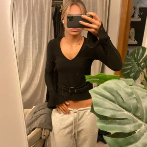 Svart ribbad topp med” bälte” - Snygg svart ribbad topp med v-ringning och ett dekorativt bälte i midjan. Toppen har långa ärmar och en figurnära passform, perfekt för en stilren outfit med extra detalj.