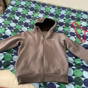 Brun hoodie från McKenzie med dragkedja - Säljer en brun hoodie från McKenzie med hel dragkedja och huva. Tröjan har långa ärmar, ribbade muddar och är i mjukt material som känns skönt mot huden. Perfekt för chill dagar eller när du vill ha en enkel och stilren look.