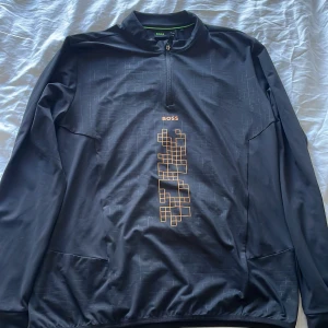 Svart BOSS half zip tröja glow print - Svart långärmad tröja från BOSS med half zip och coolt glow in the dark-tryck framtill. Tröjan har ett geometriskt mönster i orange och diskreta linjer över hela plagget. Perfekt för dig som gillar tech-vibe och snygga detaljer.