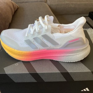 Adidas Ultraboost 5 W vit/rosa/gul - Adidas Ultraboost 5 W sneakers i vitt med gråa detaljer och en snygg färgskiftande sula i rosa och gult. Ovandel i mesh för bra andningsförmåga, klassisk snörning och bekväm Boost-sula. Perfekta för löpning eller vardagsstil.