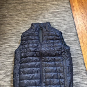 Mörkblå quiltad dunväst - Snygg mörkblå dunväst med quiltad design och hög krage. Västen har en rak passform och stängs med dragkedja framtill. Perfekt att slänga över en hoodie eller tröja när det är lite kyligt ute. Enkel och stilren look som funkar till många outfits. Storlek M.