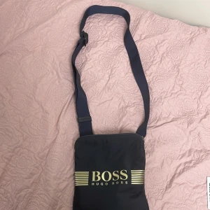 Svart axelväska från Hugo Boss - Äkta från Hugo boss butik. Snygg svart axelväska från Hugo Boss med justerbar rem och guldfärgat logotryck på framsidan. Väskan har dragkedja upptill och ett innerfack med Boss-märke. Perfekt storlek för mobil, plånbok och andra småsaker.