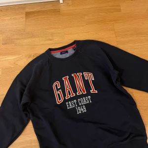 Mörkblå sweatshirt från GANT - Snygg mörkblå sweatshirt från GANT med stor rödvit logga och texten 'EAST COAST 1949' på bröstet. Tröjan har rund halsringning med röd detalj på insidan och långa ärmar. Perfekt för en chill och sportig look.
