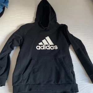 Svart Adidas hoodie med logga - Svart hoodie från Adidas med stor vit logga framtill. Klassisk känguruficka och huva. Mjuk och skön i bomullsmix, perfekt för chill eller träning. Långärmad modell med ribbade muddar för extra komfort.