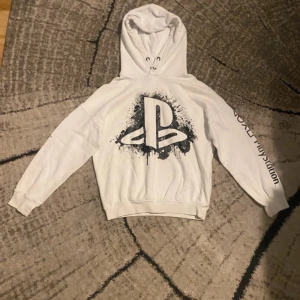 Vit PlayStation hoodie från H&M - Vit hoodie från H&M med stort PlayStation-tryck i svart på bröstet och coolt splattermönster. På ena ärmen finns PlayStation-loggan och symbolerna i svart. Tröjan har huva med snörning och ribbade muddar. Perfekt för dig som älskar gaming och streetwear.